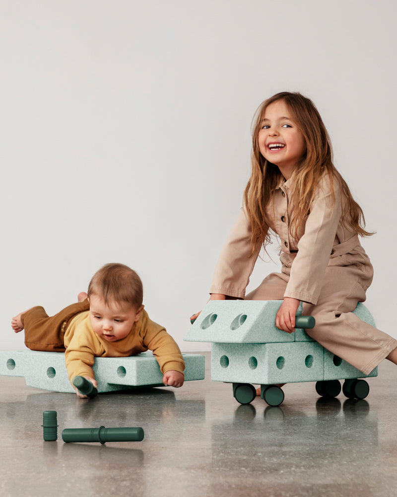 Two children enjoy active play with a ride-on toy and a rocker board build from the MODU Explorer Set.