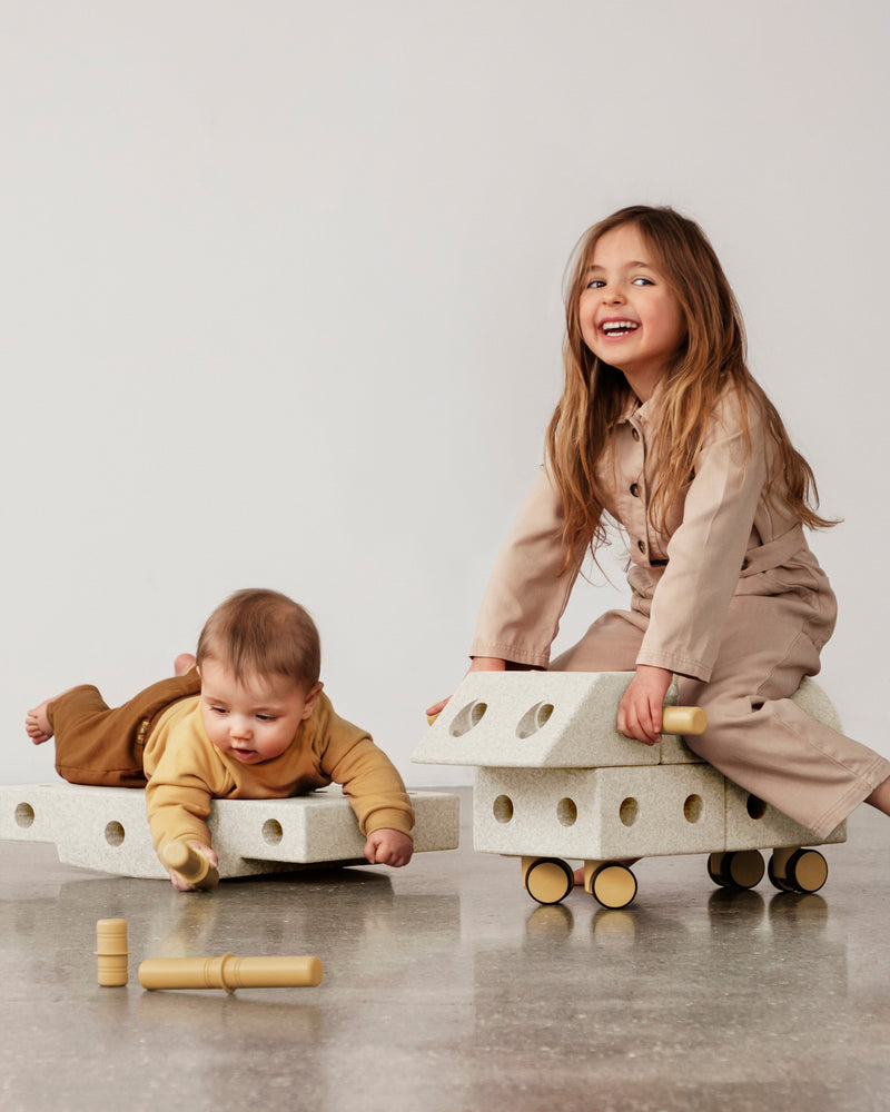 Two children enjoy active play with a ride-on toy and a rocker board build from the MODU Explorer Set.