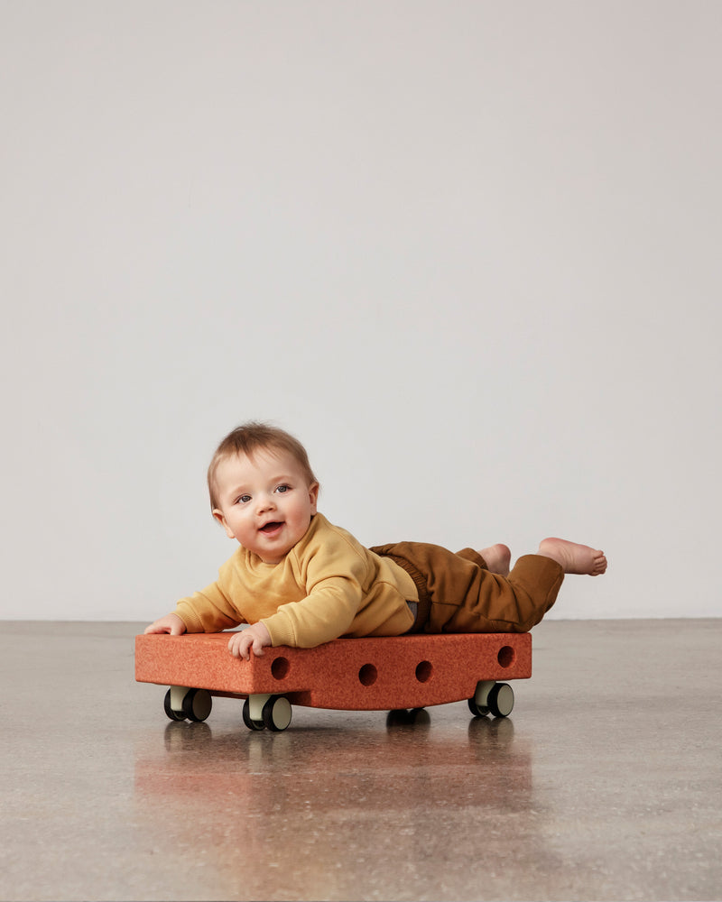 Baby playing on MODU Scooter Board in Burnt Orange Dusty Green. Encourages motor skill development.