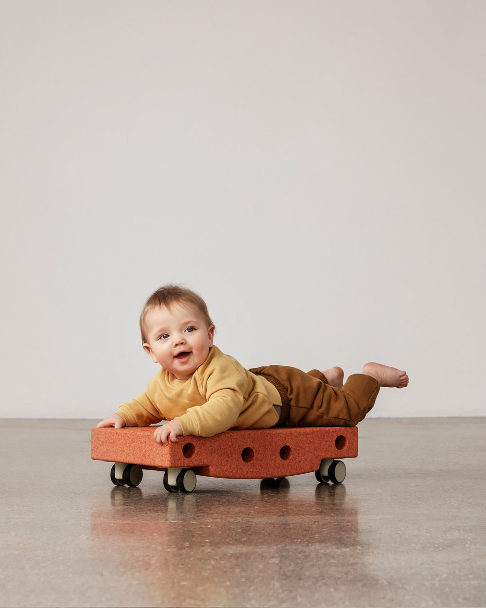 Baby playing on MODU Scooter Board in Burnt Orange Dusty Green. Encourages motor skill development.