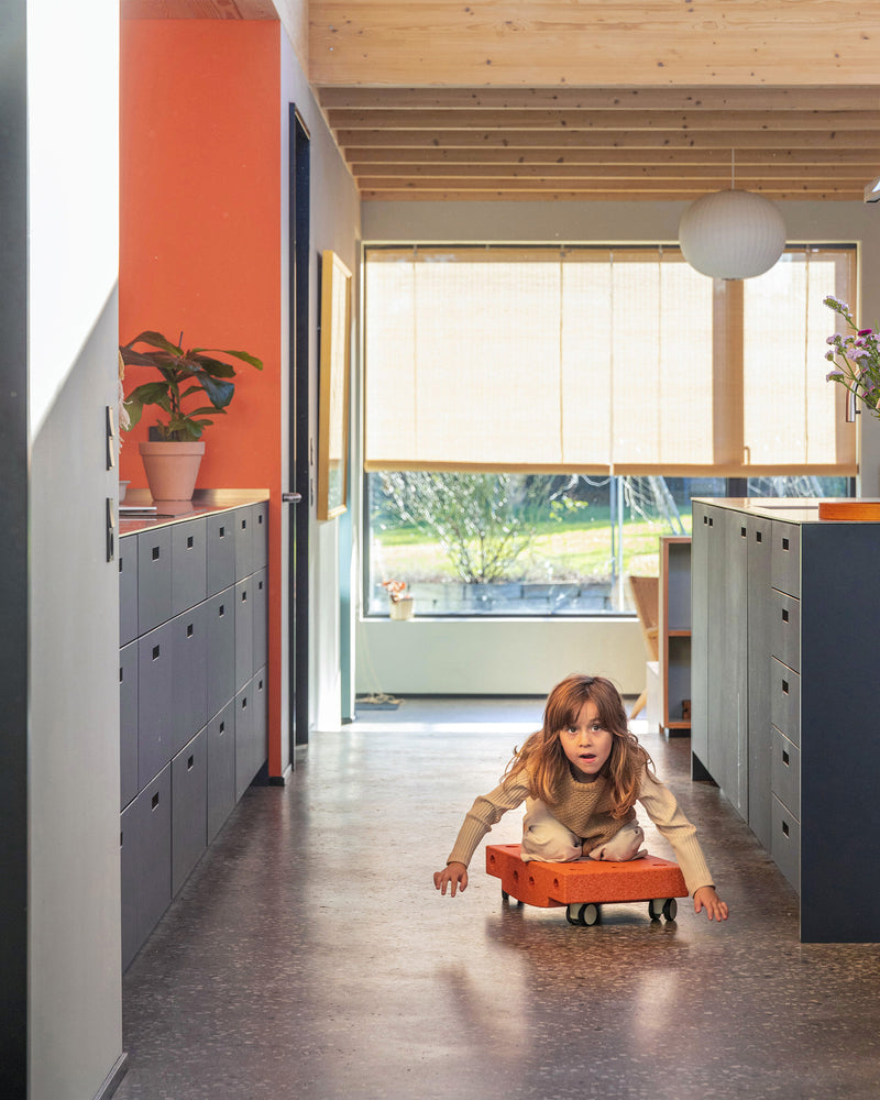 A child glides down a hallway while kneeling on a burnt orange MODU Scooter Board, practicing balance and active play at home.