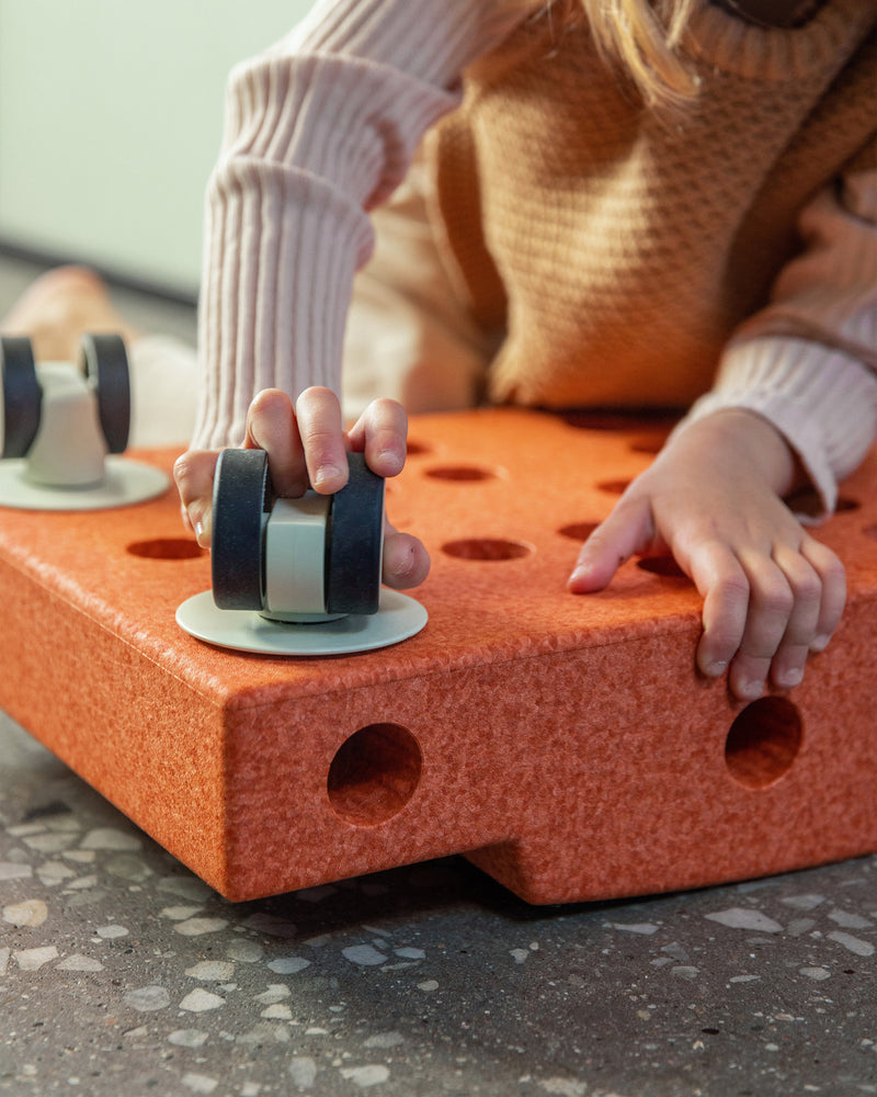 A child attaches a caster wheel to a burnt orange MODU base block, assembling their own MODU Scooter Board for active play.