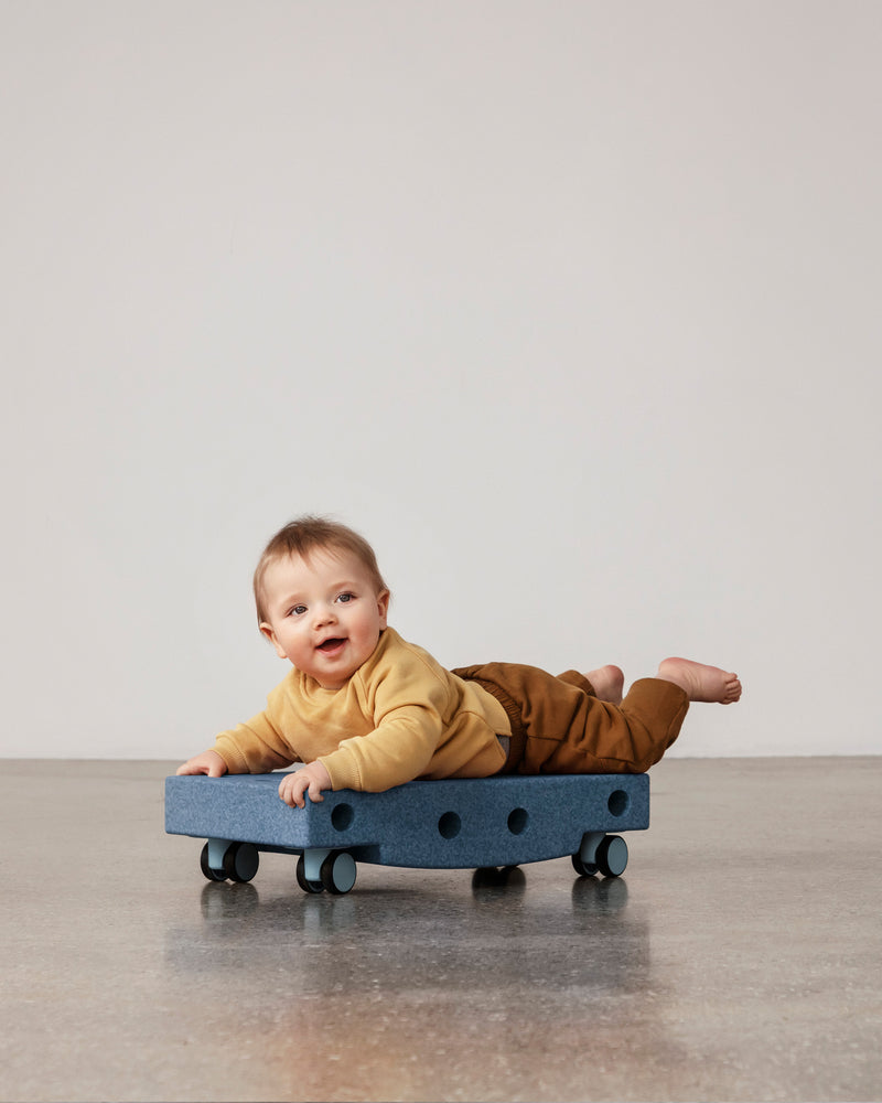A happy baby on their stomach enjoys tummy time on a deep blue MODU Scooter Board with sky blue wheels.