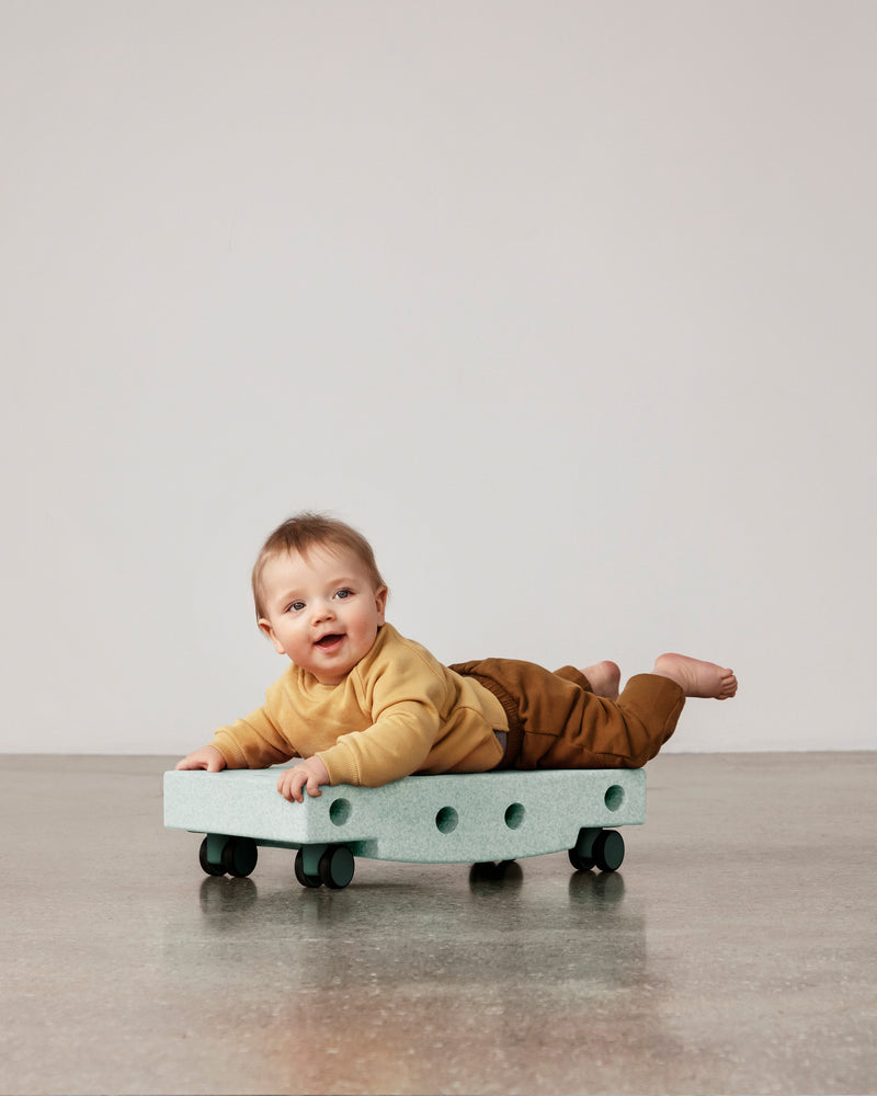 A baby lies on their stomach to practice motor skills on an ocean mint MODU Scooter Board with forest green wheels.