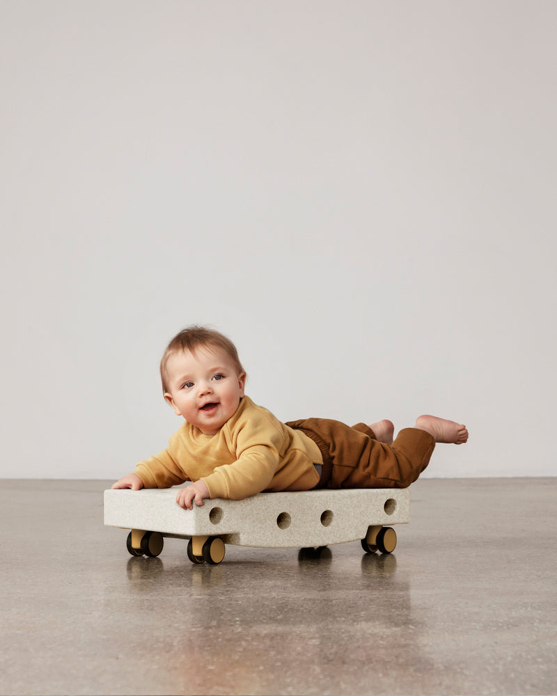 A smiling baby practices tummy time while lying on a sand grey MODU Scooter Board with honey yellow wheels.