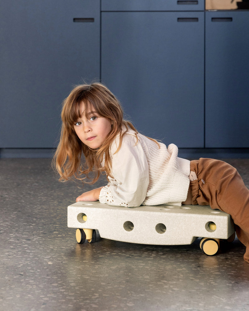A young girl lies on her stomach on a sand grey MODU Scooter Board with honey yellow wheels, showcasing an indoor toy for active play.