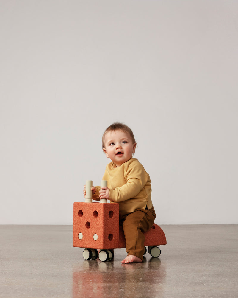 A young toddler rides a burnt orange dusty green MODU Tiny Ride.