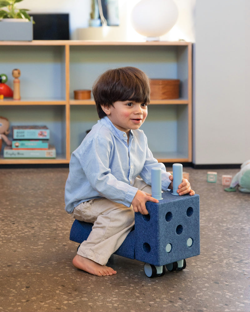 A toddler sits on a deep blue MODU Tiny Ride ride-on toy in a playroom.